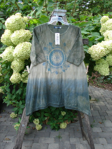 Barclay NWT Wander Tunic Tee Sunflower Mottled Vintage Camo Size 0 displayed on a wooden trellis, featuring a dip-dyed green to blue gradient and a stylized sun/mandala graphic, amidst garden greenery.