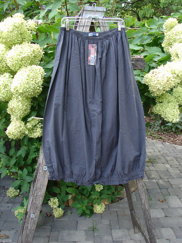 Barclay NWT Batiste Airy Lawn Skirt Unpainted Black Sand Size 0 displayed on a clear hanger, showcasing its gathered bubble style and full elastic waist, set against a backdrop of lush foliage and hydrangeas.