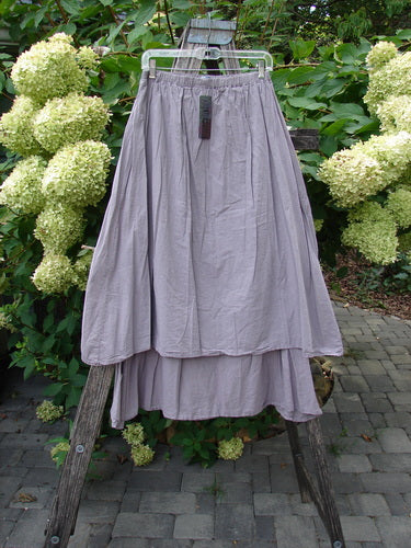 Barclay NWT Batiste Two Story Shaunting Skirt in soft mauve, features a two-tiered design and full elastic waistline, elegantly displayed on a rustic ladder amidst a lush garden backdrop.
