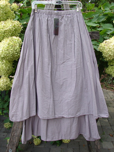 Barclay NWT Batiste Two Story Shaunting Skirt in Soft Mauve, featuring a two-tiered design with an elastic waistband, displayed on a hanger against a lush plant backdrop.