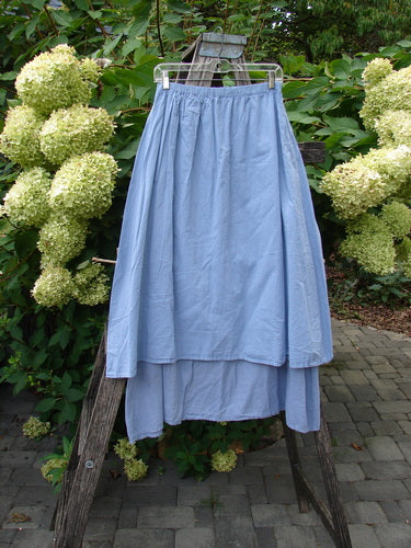 Barclay NWT Batiste Two Story Shaunting Skirt hangs on a wooden stepladder, showcasing its flowing, light-blue, double-layered design with a full elastic waist, against a garden backdrop.