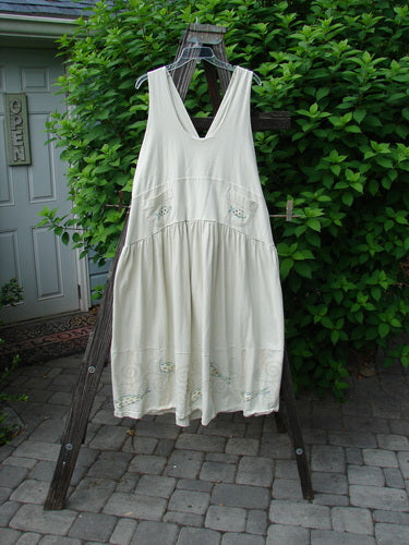 A white dress with adjustable shoulder straps, a sweeping hemline, and a criss-cross lower back. It features a double-paneled waistline, round-bottomed pockets, and a fish-themed paint design. The dress is from the 2000 Summer Collection and is made from organic cotton.