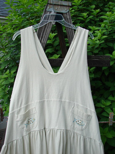 A white dress on a wooden rack, part of the 1999 NWT Tadpole Jumper Double Dot Fish collection. Made from organic cotton, it features adjustable shoulder straps, a sweeping hemline, and a criss-cross lower back. With a yoked waistline, round bottomed pockets, and fish-themed paint, this dress is a timeless piece from Blue Fish Clothing.