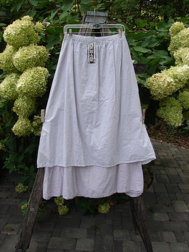 Barclay NWT Batiste Two Story Shaunting Skirt Unpainted Pearl Smoke Size 0 hanging on a clear hanger, showcasing its flowing two-tiered design and elastic waist, set against a garden backdrop.