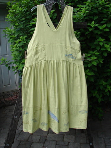 A New With Tag Tadpole Jumper dress on a clothesline. Features include adjustable shoulder straps, a sweeping hemline, and a criss-cross lower back. The dress has a yoked and empire waistline, round bottomed pockets, and a bass and moon theme paint with a blue fish patch.