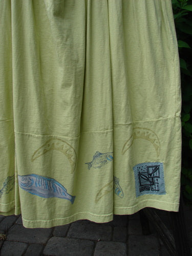 A close-up of a green dress with adjustable shoulder straps, a criss-cross lower back, and round bottomed pockets. Features a single bass and moon theme paint, and the Blue Fish patch.