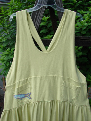 A yellow dress with a fish on it, featuring adjustable shoulder straps, a sweeping hemline, and a criss-cross lower back. It has a yoked and empire waistline, round bottomed pockets, and a single stripe pike and school theme paint around the hem. The dress is made from organic cotton and comes with the signature Blue Fish patch. Bust: 40, Waist: 50, Hips: 60, Length: 46-52 inches.