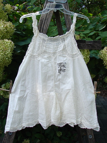 The Magnolia Pearl NWT Embroidered Eyelet Cotton Clementine Tank True OSFA features crochet lace bodice, eyelet design skirt, and Pearl Love patch, displayed outdoors on a hanger against foliage.