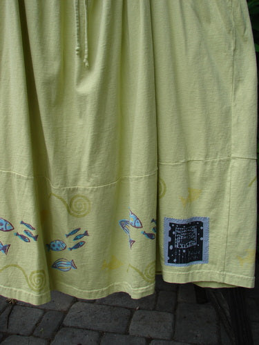 A close-up of a citron Tadpole Jumper skirt with a single stripe pike and school theme paint around the hem. Features include adjustable shoulder straps, a criss-cross lower back, and round bottomed pockets.