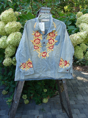 Magnolia Pearl NWT Embroidered Denim Isabel Shirt Washed Indigo OSFA, featuring red and yellow floral embroidery around the neckline and cuffs, hangs on a wooden frame, showcasing its unique distressing and signature MP patch.