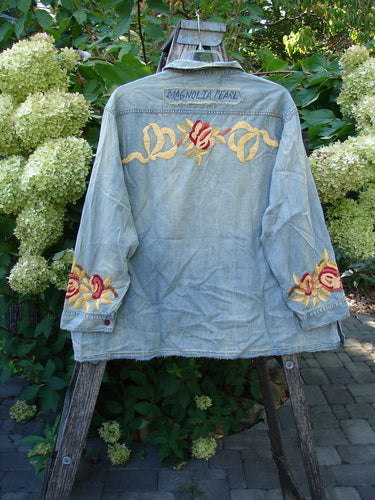 Magnolia Pearl NWT Embroidered Denim Isabel Shirt Washed Indigo OSFA displayed outdoors on a rustic ladder, showcasing intricate yellow and red floral embroidery on the back and cuffs, highlighting its artistic vintage charm.