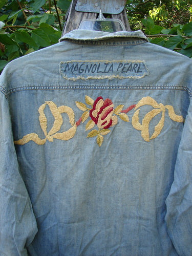 Back view of the Magnolia Pearl NWT Embroidered Denim Isabel Shirt in Washed Indigo, featuring a signature upper back patch, red rose embroidery, and distressed details, hanging outdoors.