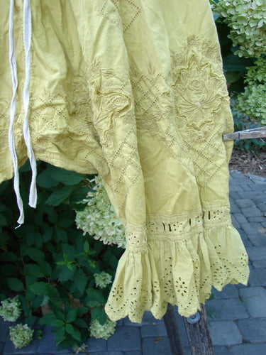 The Magnolia Pearl NWT Embroidered Khloe Bloomer Golden Daffodil OSFA features floral embroidery, eyelet lace hem, and ruffled detail, draped elegantly outdoors, highlighting its artistic craftsmanship and unique design ethos.