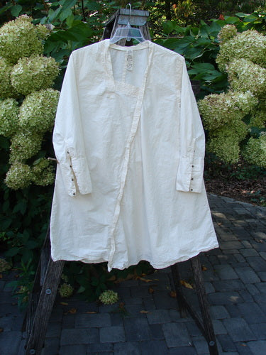Magnolia Pearl Cotton Poplin Wabi Diagonal Tunic Natural OSFA hangs on a wooden frame. The off-white tunic features unique diagonal seams, long sleeves with tiny snaps, and a slightly flared hem.