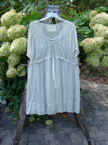 Magnolia Pearl Silk Voile Smocked Tie Back Dress in Lightest Sage OSFA displayed on a hanger, showcasing its knee-length design, three-quarter sleeves, and delicate smocking, set against lush greenery.