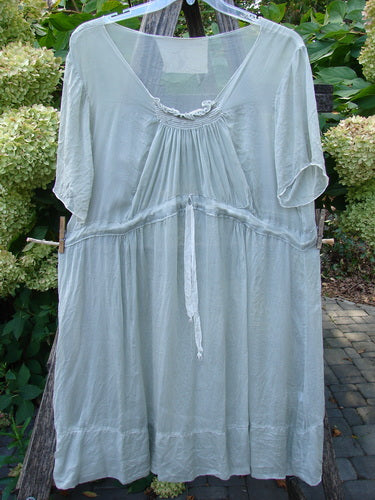 Magnolia Pearl Silk Voile Smocked Tie Back Dress Lightest Sage OSFA, featuring a loose fit, V-neckline, three-quarter sleeves, and empire waist tie, hanging outdoors amidst greenery and hydrangeas.