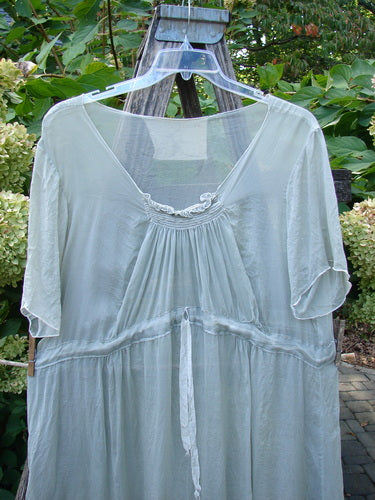 Magnolia Pearl Silk Voile Smocked Tie Back Dress Lightest Sage OSFA, featuring three-quarter sleeves, a flowing silhouette, and delicate smocking, elegantly displayed outdoors on a hanger amidst garden foliage.