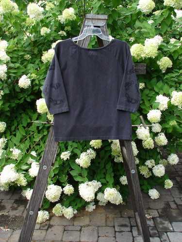 2001 Raglan Tee Top Celtic Knot Licorice Tiny Size 2: A black shirt on a wooden rack with white flowers, perfect for layering.