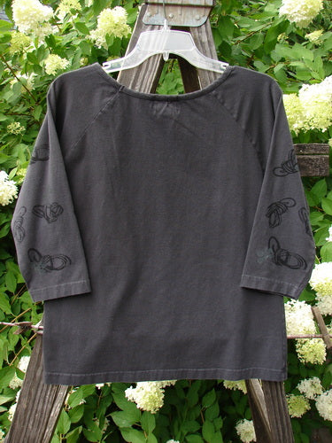 2001 Raglan Tee Top Celtic Knot Licorice Tiny Size 2: A grey shirt on a clothes rack with 3/4 length sleeves, a shallow rolled neckline, and scattered playful paint.