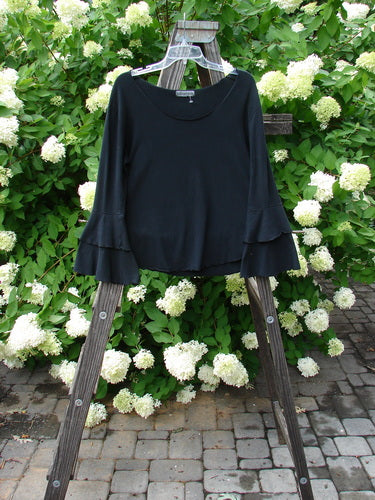 Barclay Cotton Lycra Long Sleeved Petal Top Unpainted Black Size 0: A black shirt with fancy fluttered lowers and exterior curly stitchery, on a wooden rack with white flowers.