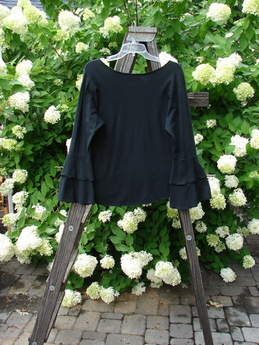 Barclay Cotton Lycra Long Sleeved Petal Top Unpainted Black Size 0: A black shirt with rounded neckline, fancy fluttered lowers, and exterior curly stitchery on a wooden easel.