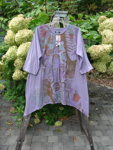 Barclay NWT Three Quarter Sleeve Color Wheel Tunic Many Moons Violet Size 0 displayed outdoors on easel, showcasing abstract graphics, lettuce-edged sleeves, A-line shape, and scoop neckline, against hydrangea backdrop.