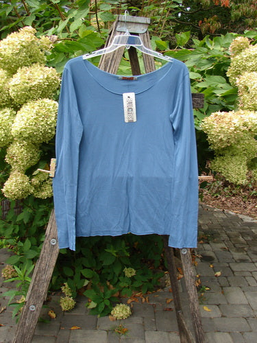 Barclay NWT Batiste Raw Edge Layering Tee Unpainted Blue Bird Size 1, featuring long sleeves, wider raw cut neckline, and rolled edgings, displayed on a hanger against a garden backdrop.