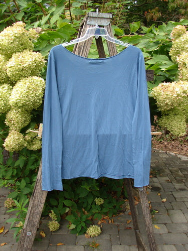 Barclay NWT Batiste Raw Edge Layering Tee Unpainted Blue Bird Size 1, displayed on a hanger over a wooden ladder, features long sleeves, a raw-cut neckline, and tapered shape, against a lush garden backdrop.