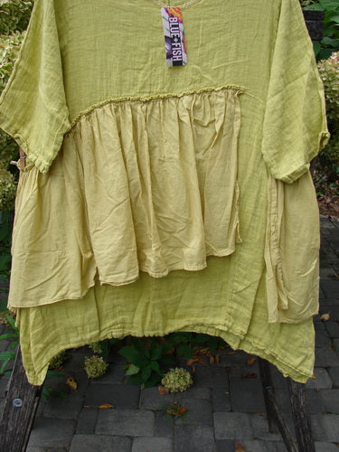 Barclay NWT Gauze Batiste Petal Urchin Flounce Dress Unpainted Citron Size 1 features a loose, layered design with ruffle detailing and an asymmetrical hem, draped over a wooden rack outdoors.