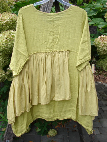 Barclay NWT Gauze Batiste Petal Urchin Flounce Dress Unpainted Citron Size 1 features a loose fit, tiered waist, and varying hemline, elegantly draped on a hanger against garden foliage.