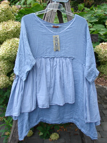 Barclay NWT Gauze Batiste Petal Urchin Flounce Dress Unpainted Dusty Sky Size 1, featuring a loose fit, scooped neckline, and three-quarter sleeves, elegantly displayed on a hanger amidst garden foliage.