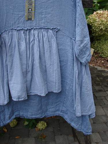 Barclay NWT Gauze Batiste Petal Urchin Flounce Dress Unpainted Dusty Sky Size 1 displayed on a wooden bench, featuring a scooped neckline, tiered skirt, and empire gathered batiste flounce.