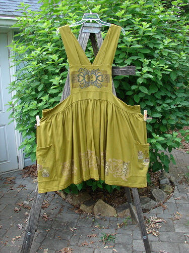 Barclay Overall Jumper Celtic Sun Glazed Tile OSFA features a creatively cut overall bib, full gathered skirt, oversized side wrap pockets, and crisscross back straps, displayed on a wooden ladder outside.