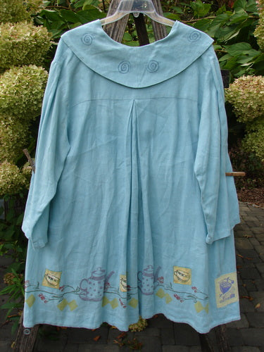 1999 Linen Breeze Jacket Tea & Cherries Spring OSFA, featuring a scalloped collar with spiral appliqués, three-quarter sleeves, and tea-time motifs, hangs on a plastic hanger against hydrangea foliage.