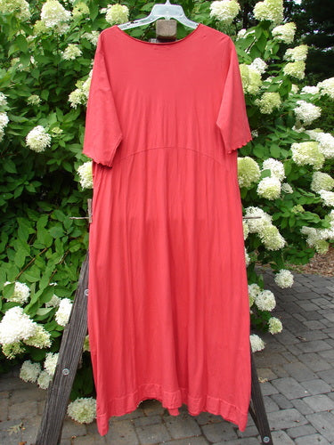 A red dress with a triangular insert, three-quarter length sleeves, and curly edgings. It has a longer narrowing shape and a single lower batiste hem flutter. The dress features exterior stitchery, deep front drop bushel pockets, and curved seams throughout. Size 2, unpainted geranium.