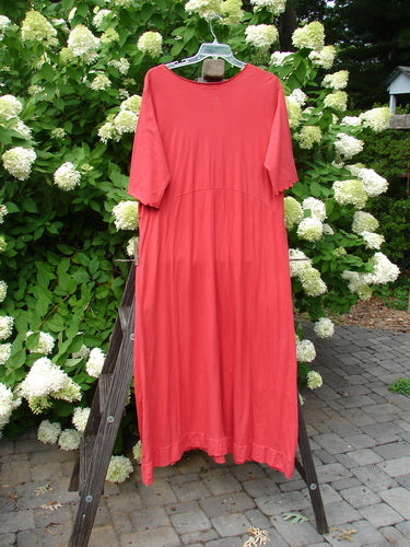 A red dress on a wooden rack, part of the Barclay Reverse Triangle Sectional Dress collection in Geranium. Features include three-quarter length sleeves, curly edgings, and a longer narrowing shape. Made from organic cotton.