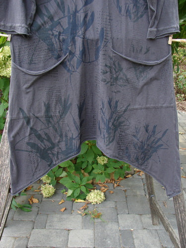 Barclay NWT Sunny Day Pocket Tunic Dress Flutter Bamboo Steel Meteorite Size 1 displayed outdoors, features an asymmetrical hem, subtle botanical pattern, and two curved front pockets, draped over a wooden frame.