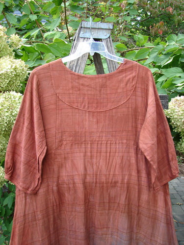 Barclay NWT Flannel Cross Over Blue Moon Pocket Dress, Terra Ombre Plaid, displayed on a hanger against a garden trellis, showcasing its loose-fitting tunic style and elbow-length sleeves.