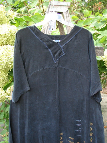 Barclay NWT Cupro Cross Over Urchin Dress, featuring a V-neck and elbow-length sleeves, hangs in a garden. The black tunic displays metallic symbols near the hem, embodying luxurious, vintage-inspired elegance.