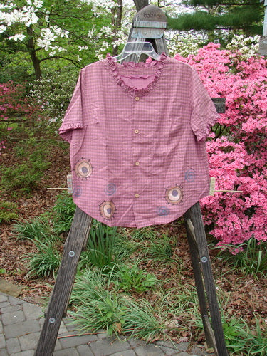 Vintage 1996 Woven Boardwalk Top featuring a gathered ruffle neck and sleeves, rounded hems, and bust line darts. Made from organic cotton. Size 1. Image: Pink shirt with frill on clothes rack, wooden ladder nearby.