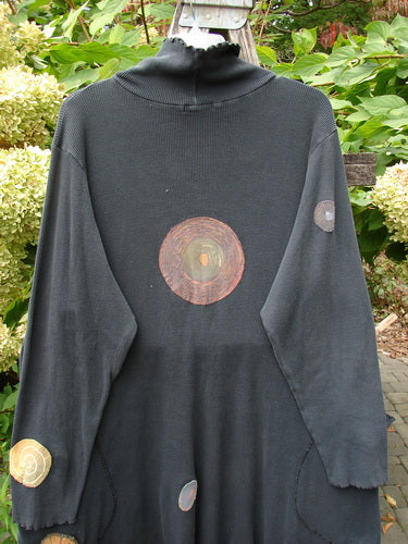 Barclay Patched Thermal Exterior Stitch Side Pocket Tunic Off Black Size 2, adorned with circular earth-tone appliqués, featuring a curl-edged turtleneck and hem, displayed against a backdrop of greenery and flowers.