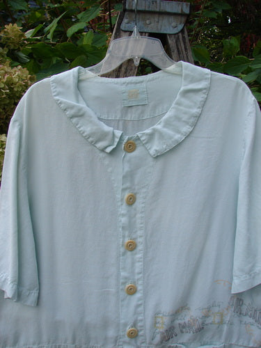 2000 Tencel Camp Shirt Airmail Shark Size 0, featuring a rounded collar, short sleeves, front drop pocket, and oversized buttons, displayed on a hanger with greenery in the background.