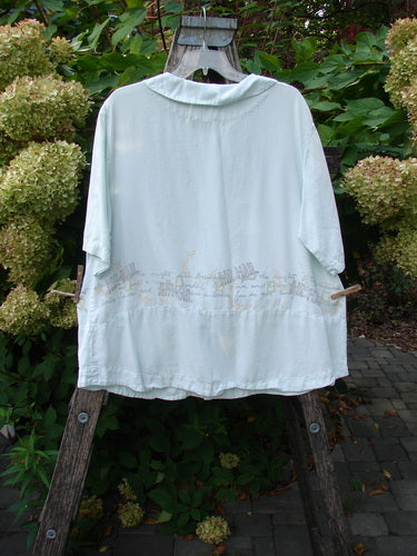 2000 Tencel Camp Shirt Airmail Shark Size 0 draped on a wooden easel, featuring a Peter Pan collar, oversized buttons, and a travel airmail theme, set against lush greenery and a brick patio.
