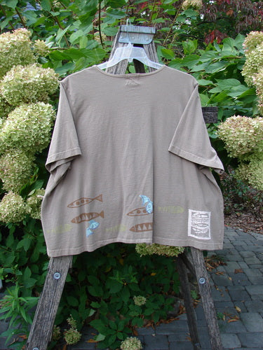1999 Tiny Pocket Top Fishie Riverbed Size 2; features a loose, cropped A-line shape with stylized fish motifs at the hem on muted olive-brown organic cotton, displayed on a wooden hanger against a natural backdrop.