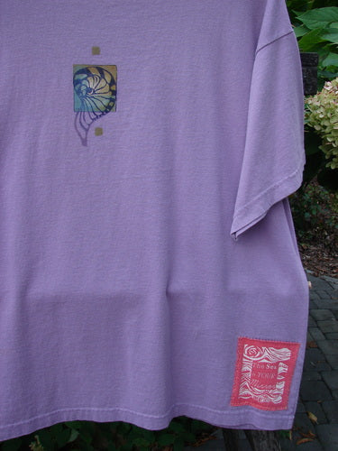 1997 Short Sleeved Tee Five Shell Jasmine Size 1: Lavender T-shirt with a multicolored spiral motif at chest and a red patch with white wavy lines near hem, laid outdoors with greenery.