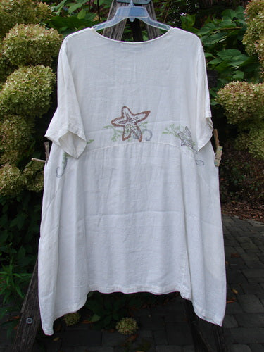 1997 Urchin Dress Sealife Froth Size 2, loose-fitting tunic with embroidered starfish motifs, displayed outdoors. The medium-weight linen showcases elbow-length sleeves and a wide neckline against a floral backdrop.