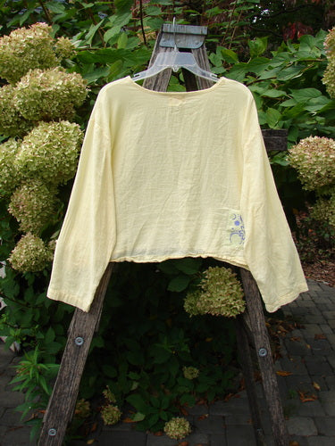 1999 Batiste Boxy Top Unpainted Buttercup Size 2, featuring a rounded neckline and billowy sleeves, hangs on a wooden stepladder, showcasing its delicate detail amidst lush greenery and hydrangeas.