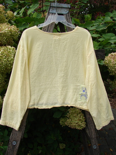 1999 Batiste Boxy Top Unpainted Buttercup Size 2, pale-yellow and billowy sleeves, displayed on a wooden ladder amidst lush greenery, embodying Bluefishfinder.com’s vintage charm.