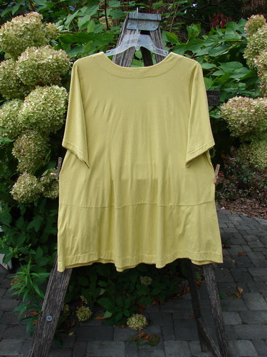 Barclay Seedling Top Unpainted Sunshine Size 2 draped over a wooden easel, showcasing its loose fit, feminine neckline, scalloped hem, and playful pockets against a serene garden backdrop.