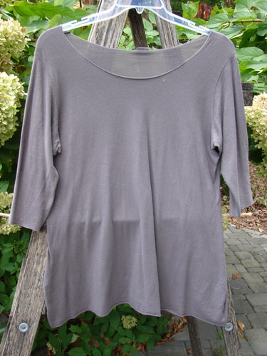 Barclay Batiste Raw Edge Layering Tee Unpainted Pebble Size 1, featuring a scoop-neck and three-quarter sleeves, displayed on a hanger outdoors against a backdrop of foliage and blooms.